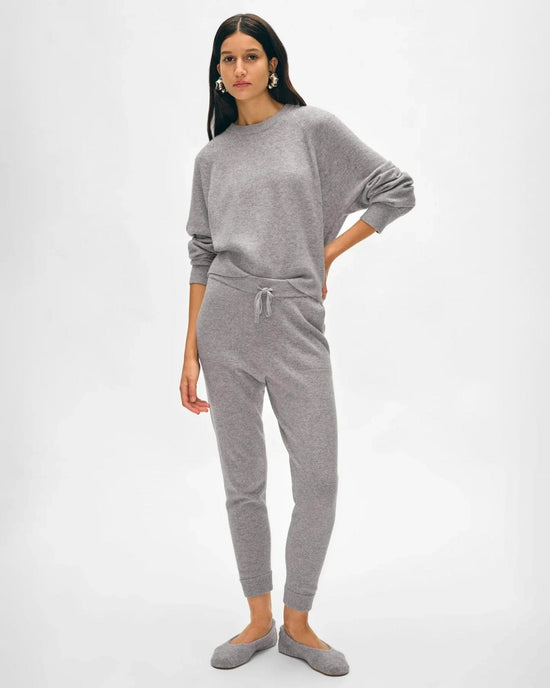 A woman with long dark hair poses calmly in the White + Warren Cashmere Jogger in Grey Heather—a cozy loungewear set with a relaxed fit, long sleeves, drawstring waist pants, and gray slippers, all against a plain background.