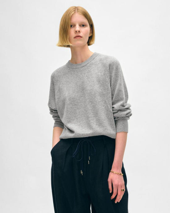 A person with straight, chin-length blonde hair wears a White + Warren Cashmere Easy Sweatshirt in Grey Heather and black drawstring pants, standing against a plain light background with hands in pockets.