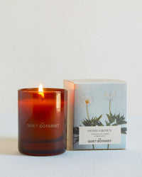 A lit Home Grown - Mini Candle by The Quiet Botanist sits beside its box, adorned with white florals and "Home Grown Tomato Leaf + Herbs" text. Enjoy this natural coconut wax candle from The Quiet Botanist.