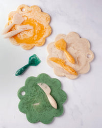 A Casa Amarosa 9in Scallop Resin Cheese/Charcuterie Board in Green, paired with a matching spatula and spreader, is artfully displayed on a white marble surface.
