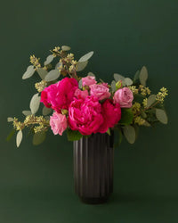 The Monroe Vase in Black by Jungle Club holds bright and light pink roses with eucalyptus leaves, set against a dark green background.
