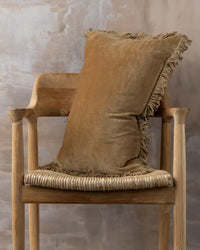 The Saarde Velvet Lumbar Pillow with Feather Insert in Nutmeg rests on a wooden chair with a woven seat, set before a textured beige wall.