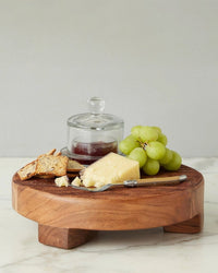 The etúHOME New Classic Round Trivet Riser, Small, made from reclaimed acacia wood, beautifully displays green grapes, crackers, cheese, a knife, and jam with a lid on white marble—a must-have kitchen serving essential.