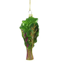 The Cody Foster Rainbow Chard Ornament is a hand-crafted decoration shaped like green chard, featuring glittery gold and purple accents, and hangs elegantly from a gold string.