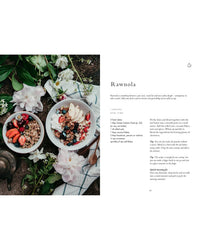 Two bowls of raw granola with fruit and coconut flakes rest on stone, surrounded by flowers; on the right, Cozy Publishing’s "Wonderful Morning - Recipes For A Delicious Start To the Day" inspires delicious breakfast ideas.