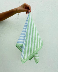 A hand holds the Casa Amarosa Stripe Hand Block Printed Scarf in Green & Blue, featuring bold vertical and diagonal stripes, set against a plain white background.