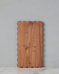 The etúHOME New Scalloped Cutting Board, Medium Rectangle Plank, features wavy edges and stands upright against a light gray wall on white marble—ideal for modern kitchen decor.