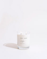 The Fern + Moss Classic 2-Wick Candle by Brooklyn Candle Studio glows in a clear glass jar against a plain, light background.
