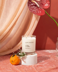The Love Potion Classic 2-Wick Candle by Brooklyn Candle Studio sits on a white pedestal with a small orange, white flowers, and a red anthurium, set against a peach drape and pink velvet for a warm, elegant look.