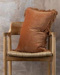 A wooden chair with a woven seat holds the Saarde Velvet Lumbar Pillow w/ Feather Insert in Terracotta, featuring fringe trim, set against a textured beige wall.