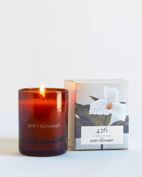 A lit 426 - Mini Candle by The Quiet Botanist, made with natural coconut wax in a brown glass jar, sits beside its box featuring a white flower illustration and the text 426, Hudson, New York, Quiet Botanist.