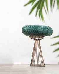 A Velvet Round Pillow in Evergreen by Casa Amarosa rests on a metal wire stool. A leafy plant appears on the right, set against a plain white background.