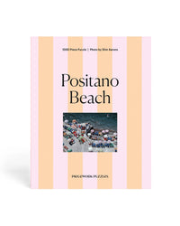 The Positano Beach - Slim Aarons Collab 1000 Piece Puzzle by Piecework Puzzles features pink and peach striped packaging with an aerial shot of a vibrant Amalfi Coast scene and the brand logo at the bottom.