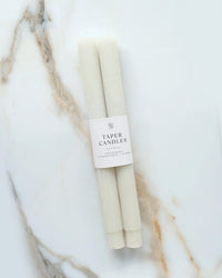 Two white unscented Neutral Taper Candles | Pillar by BOTANICA, wrapped with a label, rest on a white marble surface featuring brown and grey veining.