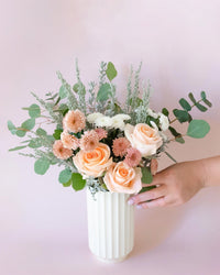 A hand touches the Jungle Club Monroe Vase in Vintage White, filled with peach roses, pink chrysanthemums, white flowers, and assorted greenery against a soft pink background.