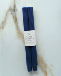 Two unscented Navy Taper Candles | Fluted by BOTANICA are bundled with a white label reading "Taper Candles" and displayed elegantly on a white and brown marbled surface.