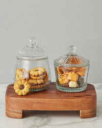 Two glass jars with lids rest on the etúHOME New Classic Rectangle Trivet Riser, Small—one jar holds assorted cookies, including a flower-shaped treat; the other contains wrapped caramel candies—all set against a neutral gray background.