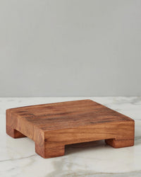 The etúHOME New Classic Square Trivet Riser, Small, handcrafted from reclaimed acacia wood with four short legs, rests on a white marble surface against a plain gray background.