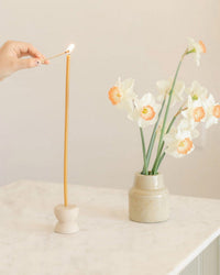A hand lights a tall candle in The Floral Society's Gem Petite Long Stem Candleholder in Sand on light marble. Next to it, a beige vase with white and orange daffodils adds charm to this modern decor scene with a soft, blurred background.