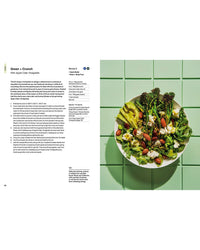 A bowl of salad from The Salad Project by Random House, featuring broccolini, avocado, almonds, feta, and greens, sits on green tiles. To the left is a printed recipe for Green + Crunch with Apple Cider Vinaigrette.