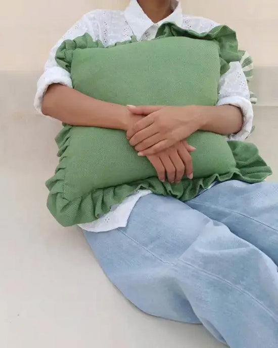 A person in a white eyelet shirt and light blue jeans sits with arms crossed over a Casa Amarosa Slate Green Ruffle 16x16in Pillow in their lap. Their face is not visible.