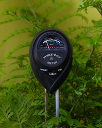 The Jungle Club 3-in-1 Moisture Meter in Black is placed among green ferns, showing moisture, light, and pH levels. The dial reads "moist," indicating healthy plants against a vibrant yellow-green background.