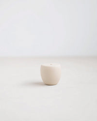 The Bulb Petite Long Stem Candleholder in Sand by The Floral Society is a small, round, off-white ceramic piece with a tiny hole on top, centered on a light surface with a minimal, soft background.