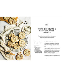 A wire rack with white chocolate and salty liquorice cookies, inspired by Nordic food, sits beside a small bowl of chocolate. Nearby are more cookies and a gold spoon. The “Cookbook: Crumbs of Joy” by Cozy Publishing is shown on the right.