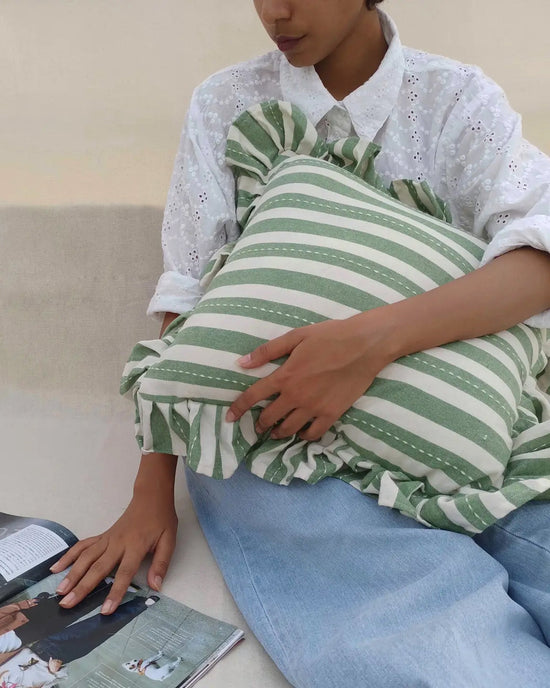 A person in a white eyelet blouse and blue jeans holds the Casa Amarosa Striped Green Ruffle 16x16in Pillow, made from 100% cotton, while resting their hand on an open magazine.