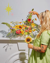 At Baske’s Thanksgiving Floral Workshop, a woman arranges a vibrant flower display in a white vase, holding a sunflower. She wears a light green dress; pastel star decorations adorn the wall in the background.