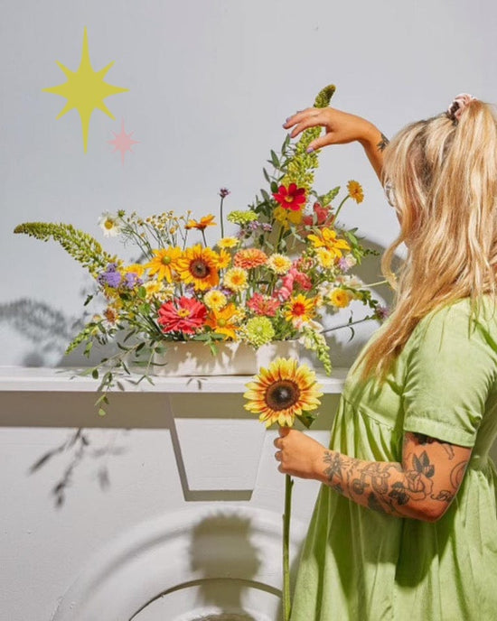 At Baske’s Thanksgiving Floral Workshop, a woman arranges a vibrant flower display in a white vase, holding a sunflower. She wears a light green dress; pastel star decorations adorn the wall in the background.
