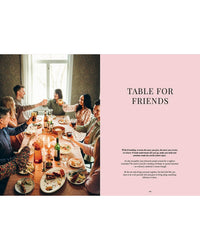 Six friends gather around a table enjoying Nordic dishes from "Cookbook: Crumbs of Joy" by Cozy Publishing, toasting amid candles and drinks. On the right, a pink page reads “TABLE FOR FRIENDS” with text below.