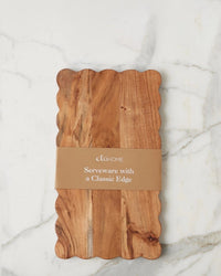 The etúHOME New Scalloped Cutting Board, Medium Rectangle Plank, features scalloped edges and rests on marble. A brown paper band around the board reads etúHOME Serveware—perfect for modern kitchen decor.