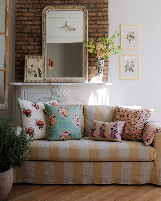 A striped sofa with a Soil to Studio Zeenat Linen Pillow (14 x 20) sits before a white mantle topped with a large mirror, botanical prints, and flowers. In the foreground is a potted plant.