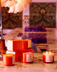 Three lit ROEN La Condesa Candles and two matching boxes, made from coconut soy wax and premium fragrance oils, sit on a marble countertop with a decorative lamp and patterned tiles softly glowing in the background.