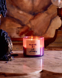 A lit Nueve Leones Candle by ROEN, made from a coconut soy wax blend, glows in a pink and purple glass jar on marble, melted wax drips down the side as a decorative lamp adds to the warm, cozy ambiance in the background.