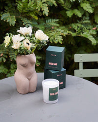 A curvy vase with white flowers, a ROEN Hotel Flori Candle in a glass, and two green candle boxes rest on a round gray outdoor table surrounded by leafy greenery and a green chair.