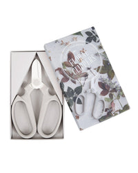 A pair of Japanese Floral Clippers in Fog by The Floral Society rests in a box beside its lid decorated with botanical illustrations and labeled “Floral Clippers.”.