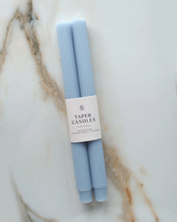 A pair of fluted Cornflower Blue Taper Candles by BOTANICA are bundled with a white "Taper Candles" band and displayed on veined marble. These unscented candles add an elegant touch to any space.