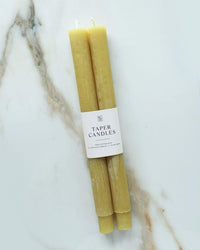 Two Martini Olive Taper Candles by BOTANICA, featuring a white "Taper Candles" label, rest on a white and marbled surface.