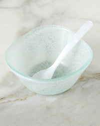 A translucent, frosted glass bowl rests on marble, complemented by the etúHOME Acrylic Dipping Spoon in Pearl—an elegant accessory perfect for entertaining at your next gathering.