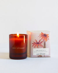 The Quiet Botanist Orchard Sun - Mini Candle, made with natural coconut wax, sits beside its box decorated with orange flower designs and bold "Orchard Sun" text, adding a botanical touch to your space.