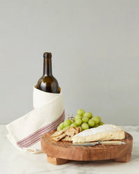 A bottle of wine in a white cloth, cheese with a knife, crackers, and green grapes displayed on the etúHOME New Classic Round Trivet Riser, Medium, all atop a marble surface.
