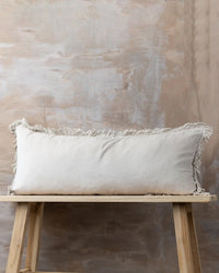 The Saarde Velvet Body Pillow w/ Feather Insert in Clay rests on a light wooden bench, set against a textured beige and gray wall.
