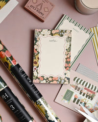 A flat lay of floral and striped stationery—featuring Amy Heitman’s Flower Market Note Pad with tear-off sheets, plus notepads, wrapping paper, cards, and a pastel candle—all arranged on a pink surface in soft decorative patterns.