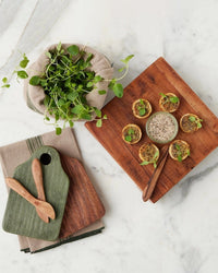 A wooden board with mini tarts and dip sits on marble next to the etúHOME New Classic Square Trivet Riser, Small—stacked and crafted from reclaimed acacia wood—with slender wooden utensils and fresh leafy greens nearby.
