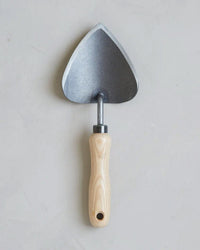 The Floral Society Potting Trowel features a wide, triangular boron steel blade and a smooth, light wooden handle—an essential garden tool, shown against a plain light gray background.