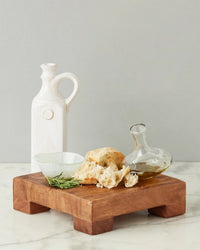 The etúHOME New Classic Square Trivet Riser, Small, displays torn bread, a glass bowl, a carafe of olive oil, rosemary sprigs, and a white ceramic oil bottle on marble with a gray background.