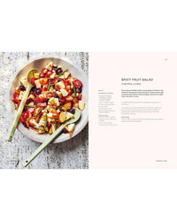 A vibrant spicy fruit salad with mixed fruits, nuts, and herbs sits on a textured white surface with two serving spoons inside, alongside the "Thai Made Easy" cookbook by Chronicle Books displayed to the right.