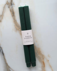 Two fluted green taper candles from BOTANICA, labeled "Evergreen Taper Candles | Fluted," rest on a white surface with brown and gray veining. Unscented, they add an elegant touch to any decor.